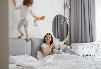 Obraz na płótnie Canvas Mama with two kids at home in bedroom. Baby infant resting on mothers hands, while older sister playing, action, jumping on the bed near, Cuddle a baby while talking to toddler daughter