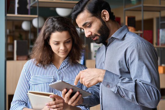 Diverse Friendly Coworkers Colleagues Indian Male Manager And Latin Female Employee Talking In Office Space Using Tablet Device. Effective Management, Teaching And Mentoring New Employee Concept.