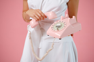 Female hands with a pink wired phone close up on a pastel pink color background. Woman in a white dress is holding phone. Business ..lady with the telephone.
