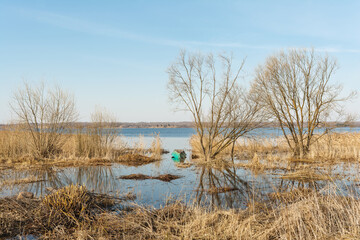 Spring flood blue lake. Trees still without leaves grow in a meadow flooded with water. An overturned wooden boat in the water on the shore of a lake covered with dry grass