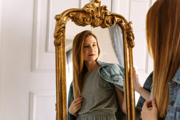 Cozy home atmosphere. Stylish redhead woman in gray dress and jeans sits on a chair at home and looks in the mirror. Portrait beautiful young woman happy smile relax in living room interior