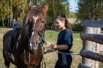 Cute girl gets ready for a ride on a horse and saddles it on an autumn day