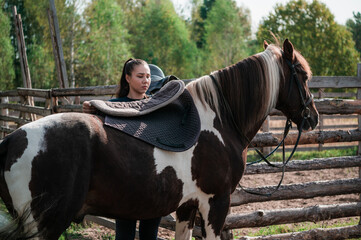 Cute girl gets ready for a ride on a horse and saddles it on an autumn day