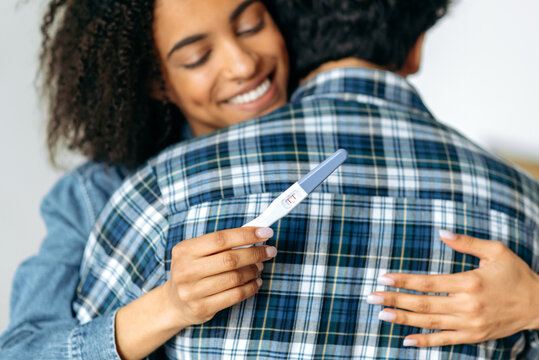 Positive pregnancy test concept. Happy african american young woman hugs her beloved husband, holding a positive pregnancy test, happy about good news, preparing to become a mother, happy family