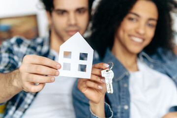 New home owners. Happy young couple holding paper house and keys, sitting on a sofa in their new modern apartment or house. Defocused african american girl and hispanic guy happy to buy their own home