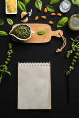 Basil pesto sauce on a small wooden table with basil leaves, parmesan cheese, almonds, garlic, olive oil and salt as ingredients, and a notebook for copy space on a black background, vertical image.