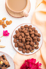 Almond in chocolate dragees on ceramic plate and a cup of coffee on white concrete background. Top view, close up.