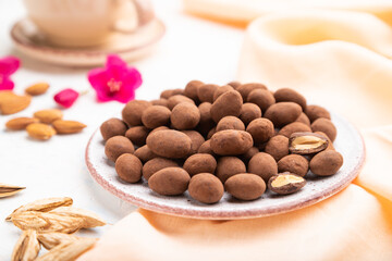 Almond in chocolate dragees on ceramic plate and a cup of coffee on white concrete background. Side view, selective focus.