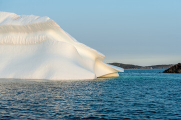 A large white iceberg formation floating in the cold ocean with layers of textured ice and snow. The ice is in transition melting from the warm rays of the sun at sunset. The berg is a blocky shape.