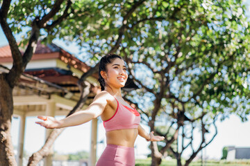 Attractive and fit young smiling Asian woman practicing yoga with stretched arms in outdoor park during morning routine for a healthy body