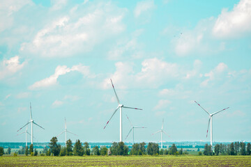 many windmills stand on a large field spinning in the day against a sunset orange sky. Alternative...