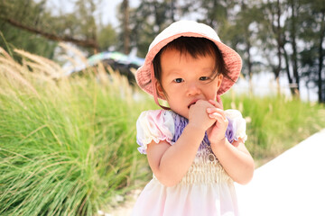 A little cute adorable embarrassed and shy asian baby girl of​ 1 - 1.5​ years​ old​ wear pink hat and pastel dress in the green garden, putting her finger in her mouth.