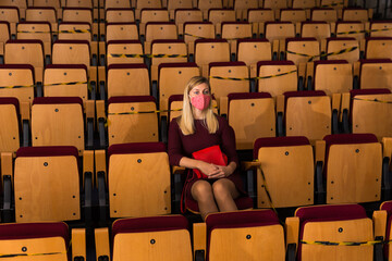 Young woman theatergoer wearing protective mask sitting in empty theater hall, watching...