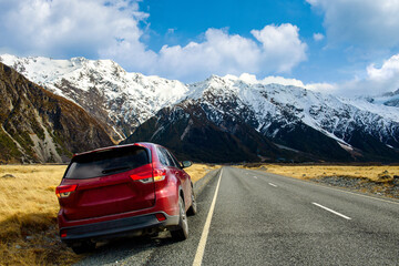 Red SUV car on the roadside, background is big and tall Mountains in winter, dry grass turns yellow in blue skies and clouds in the daytime in Mount cook  National Park in southern island, New Zealand