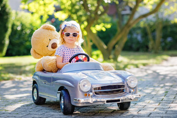 Little adorable toddler girl driving big vintage toy car and having fun with playing with plush toy...