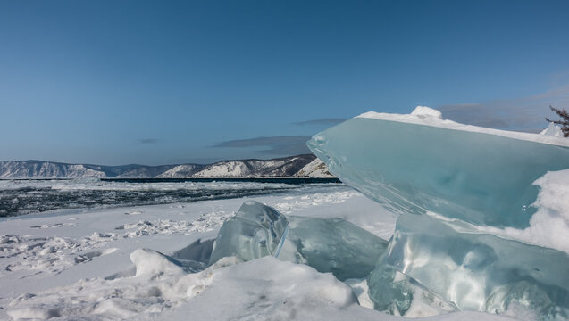 Turquoise ice hummocks on the snow-covered shore. There are melted ice floes on the river. Close-up. Background - blue sky, mountain range.