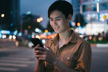 Young Asian man is using his phone while walking in the street at night