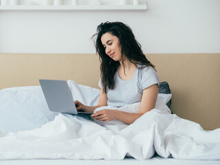 Morning chat. Online communication. Modern technology. Home leisure. Relaxed happy woman enjoying reading message using laptop in bed in light bedroom.