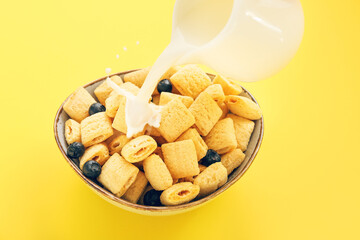 Pouring of milk into bowl with tasty corn pillows and blueberries on color background