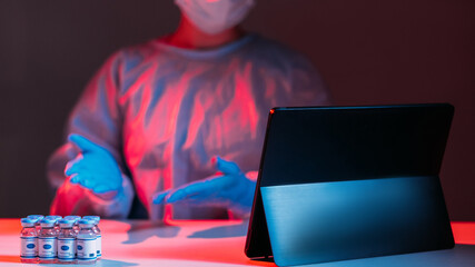 Covid-19 vaccine. Remote consultation. Pandemic telemedicine. Female doctor in ppe demonstrating vial dose on tablet online in red blue neon light in dark laboratory.