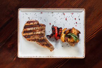 Meat Steak on a white ceramic platter with fried potatoes, vegetables, spices and herbs on a wooden table. Top view, horizontal orientation, no people.