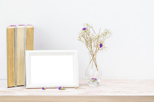 An empty mock-up of a wooden picture frame next to a stack of old books. A vase of dried white and purple flowers on a wooden table. Interior and home office design in the Scandinavian style.