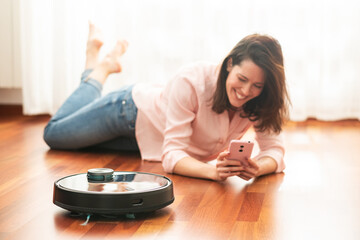 Young caucasian woman smiling in the living room and using automatic vacuum cleaner to clean the floor, controlling smart machine housework robot with smart phone at home