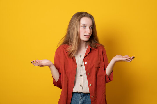 Cute Puzzled Teen Girl, Spreads Hands With Doubt, Dressed In Orange Shirt, Isolated Over Yellow Background. Woman Faces Dilemma, Being Unsure.