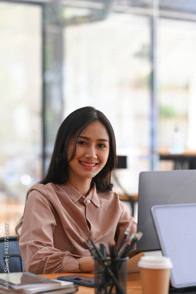 Wall mural portrait of happy young female employee siting at her workplace and smiling to camera.