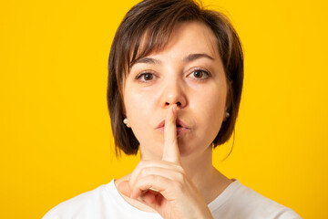 Serious young female dressed casually holding finger at her lips asking not to make noise or hold tongue and conceal some private confidential information. Top secret.
