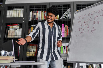 Smiling Indian teacher during online lesson, online learning, distance learning, he writes on the board, looks at the computer