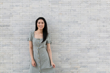 Portrait of a happy Asian girl in dress standing by the stone wall. Young traveling woman standing near stone wall during holiday. Tourist woman standing by the wall.