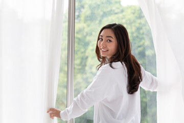 Young woman stand open white curtains at the window in morning after waking up in bedroom. Happy female opening window curtains at home. Woman standing by bedroom window opening curtains.