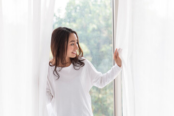 Young woman stand open white curtains at the window in morning after waking up in bedroom. Happy female opening window curtains at home. Woman standing by bedroom window opening curtains.