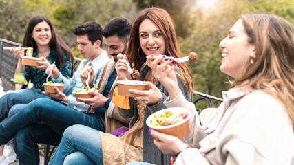 Gathering of young people eating take away organic food in plastic-free bowls and cutlery together  in the park. Focus on the second from right smiling and talking. - obrazy, fototapety, plakaty