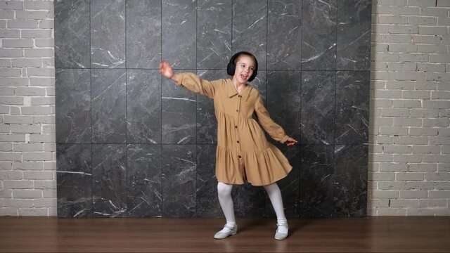 Delighted active girl in brown dress dances wearing black headphones against designed marble wall surrounded by grey bricks in hallway