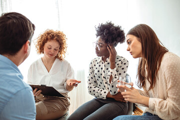 A diverse group of adults sit in a circle in their chairs and listen to and sympathize with each...