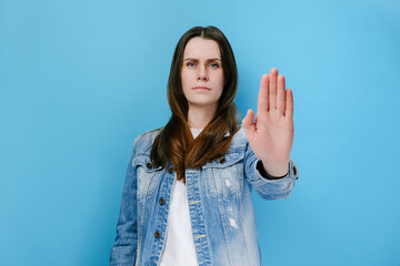 Time to stop. Young angry woman extends palm at camera, prevents you from making mistake, makes forbid sign, smirks face, restricts or warns, wears denim jacket, isolated on blue studio background