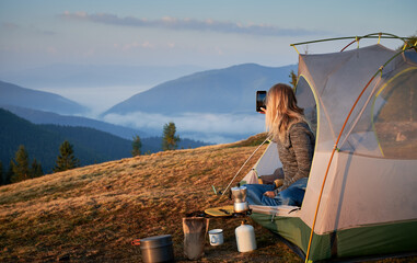 Rear view of girl photographing morning landscape with smartphone from her tent set up on hill during breakfast outdoors. Concept of active camping in the mountains, tourist equipment and photography.