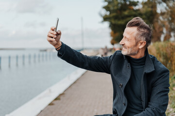 Man seated at the waterfront on a cold winters day taking his selfie