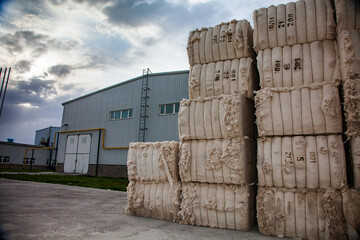 Purifying cotton plant. Stack of cotton packs (right) and industrial building left. Sky with clouds and sun.