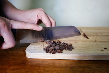 A closeup picture of a woman mincing pieces of dark chocolate into small pieces using a knife and a wooden cutting board before adding it to her cookie dough before baking.