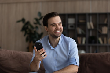 Obraz na płótnie Canvas Smiling young Caucasian male user or client text message online on smartphone look in distance thinking pondering. Happy man relax at home use cellphone gadget make plan. Communication concept.