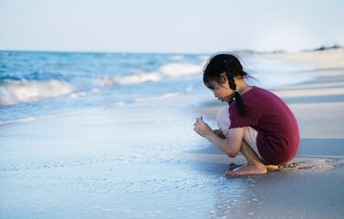 A cute young Asian girl, sitting on a white sandy beach with calm warm water, collecting seashells while on vacation with her family.