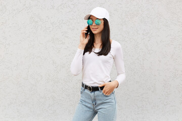 Confident attractive woman wearing baseball cap, casual shirt, jeans and sunglasses, talking via cell phone, dark haired female using smart phone against white wall.