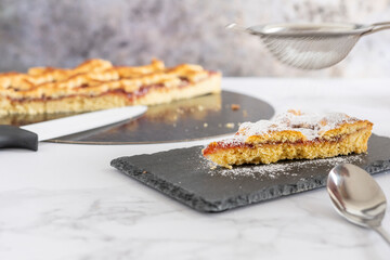 Putting icing sugar on a piece of berry jam cake on a black rectangular plate, Jam cake out of focus next to a knife. Marbled background