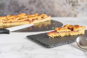 Piece of forest fruit jam tart on a black rectangular plate, out of focus jam tart with a knife. Marbled background