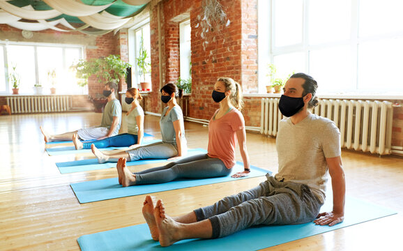 fitness, epidemic and health care concept - group of people wearing face protective black masks for protection from virus disease doing yoga seated staff pose on mats at studio