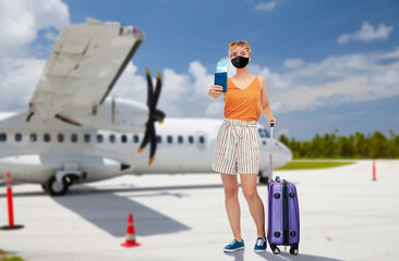 tourism, vacation and people concept - happy teenage girl or young woman wearing face protective black mask with travel bag, passport and air ticket over plain at airport on background