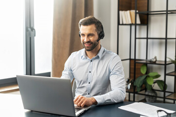 Cheerful mature fair-haired man freelancer in headset working from home during pandemic, having video chat with employer or clients, using modern laptop and documents, copy space, typing on keyboard